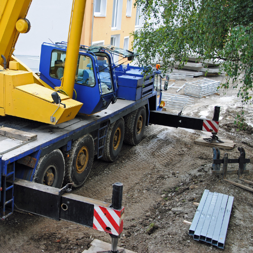 Crane Rental ServicesYellow and blue crane on a flatbed truck at a construction site with buildings and trees in the background.
