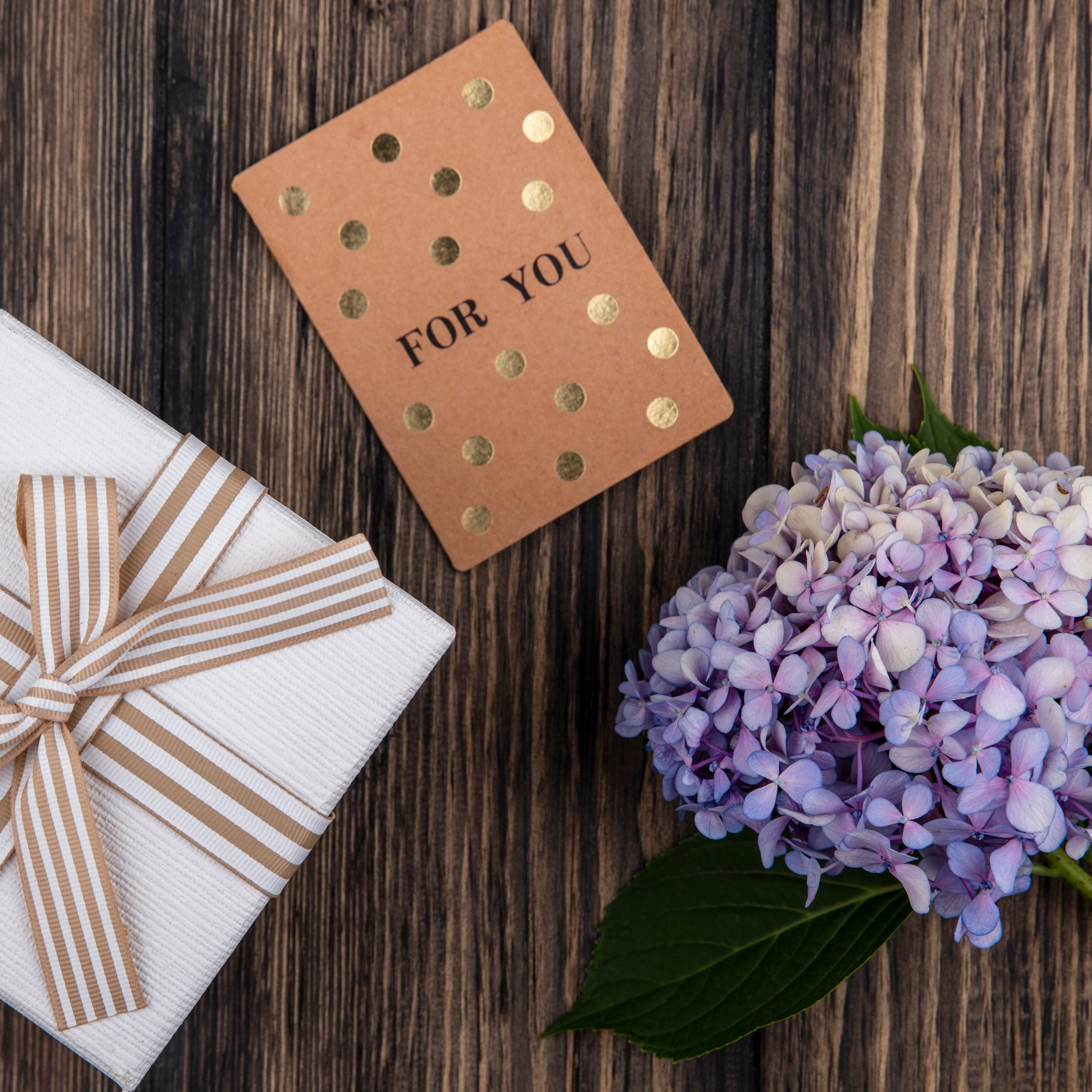 Gift box with striped ribbon, 'For You' card, and purple flowers on a wooden surface